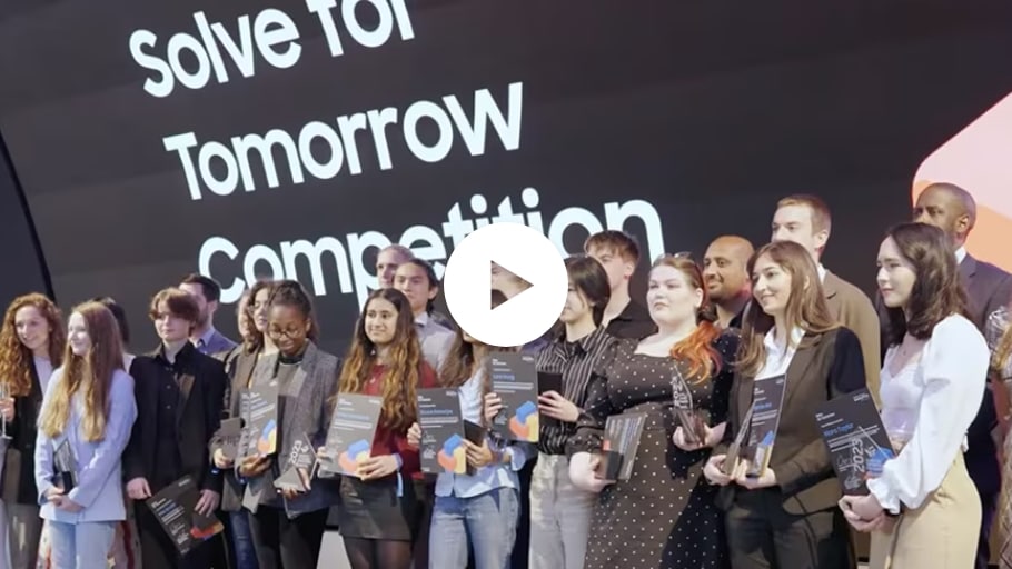 A range of youths stand in front of a sign that reads, ‘Solve for Tomorrow Competition’. Some are holding certificates. A play button in the middle shows that this is a video ready to be played. 
