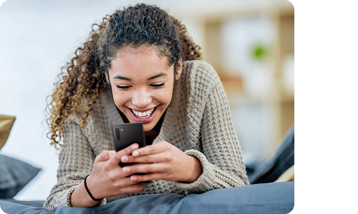 A woman leaning on a couch, smiling and watching her smartphone.