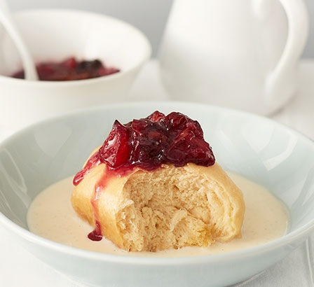 There is a dumpling in a white plate, and roasted plum compote on its top. And vanilla custard drizzled around the dumpling.