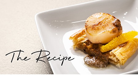 At the center of a white square plate, there is a slice of Pissaladière topped with a slice of orange and pan-grilled scallop. Over the scallop, there is a drizzle of olive and orange puree to complete the simple but elegant plating.