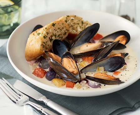 Steamed mussels and roasted rainbow roots paired with white garlic bread on it are in an white bowl.