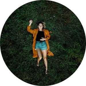 A moody drone shot of Iz Harris laying on a bed of dark green grass, she is wearing a tan jacket, black top, denim jeans and black sandals.