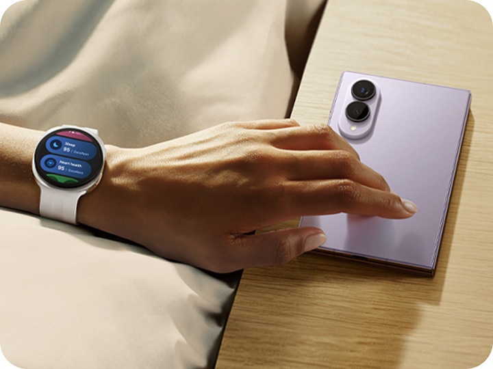 Galaxy Z Fold8 in Lavender is folded and screen down on a surface. A hand is reaching out with Galaxy Watch9 in Cream on the wrist, showing the sleep score and heart health score