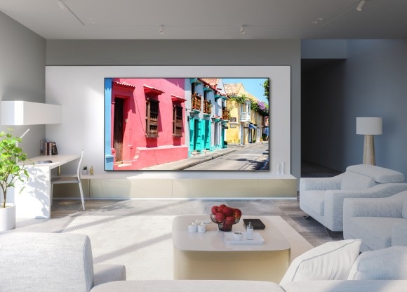 A 98 inch Supersize TV is mounted on a wall in a living room displaying colorful buildings.