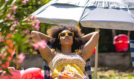 A woman with sunglasses relaxes in her backyard, surrounded by a beach umbrella, inflatable duck and lawn chair.