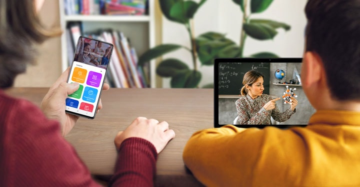 Вид женщины через плечо, держащей в левой руке смартфон Galaxy A51, который отображает уроки Дженни в четвертом классе. Рядом с ней сидит мальчик, наблюдая за тем, как учительница ведет урок естествознания, держа в руках молекулярную модель.