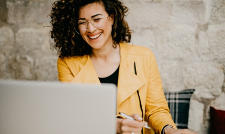 A curly-haired woman laughs while video chatting on her Galaxy laptop. She is wearing a mustard yellow jacket and holding a pen in her right hand. She is sitting in front of a beige cemented wall.