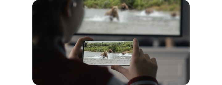 A young girl is holding a Galaxy Note10 in her both hands. And there is a video of bears in the water scene on her phone that is also on the TV in front of her