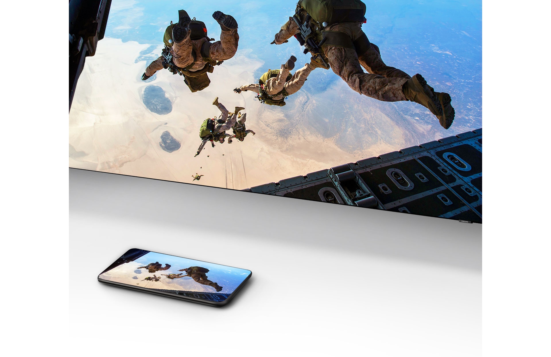 Видео, изображающее людей, прыгающих с парашютом, воспроизводится на смартфоне перед телевизором Samsung Smart TV, на экране которого дублируется тот же контент.