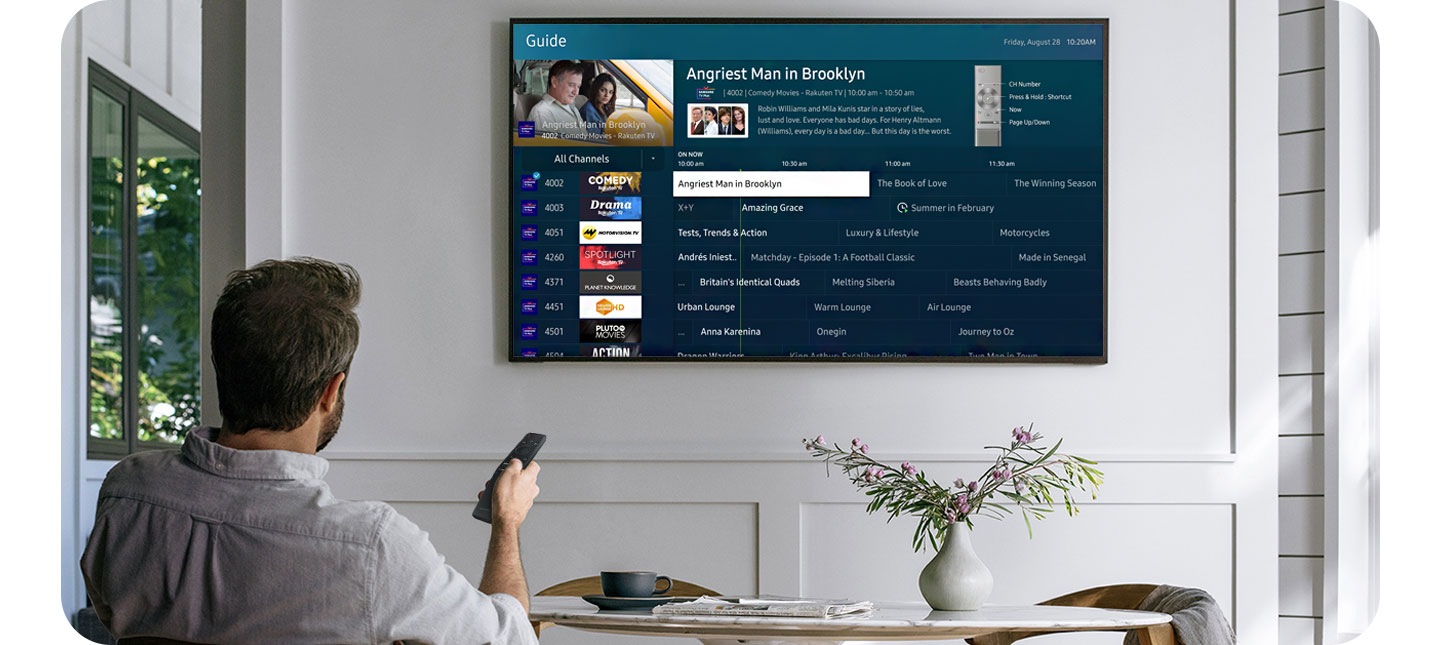 Мужчина направляет универсальный пульт One Remote на Samsung Smart TV, висящий на стене. Мужчина листает список каналов Samsung TV Plus, отображенный на экране Samsung Smart TV.