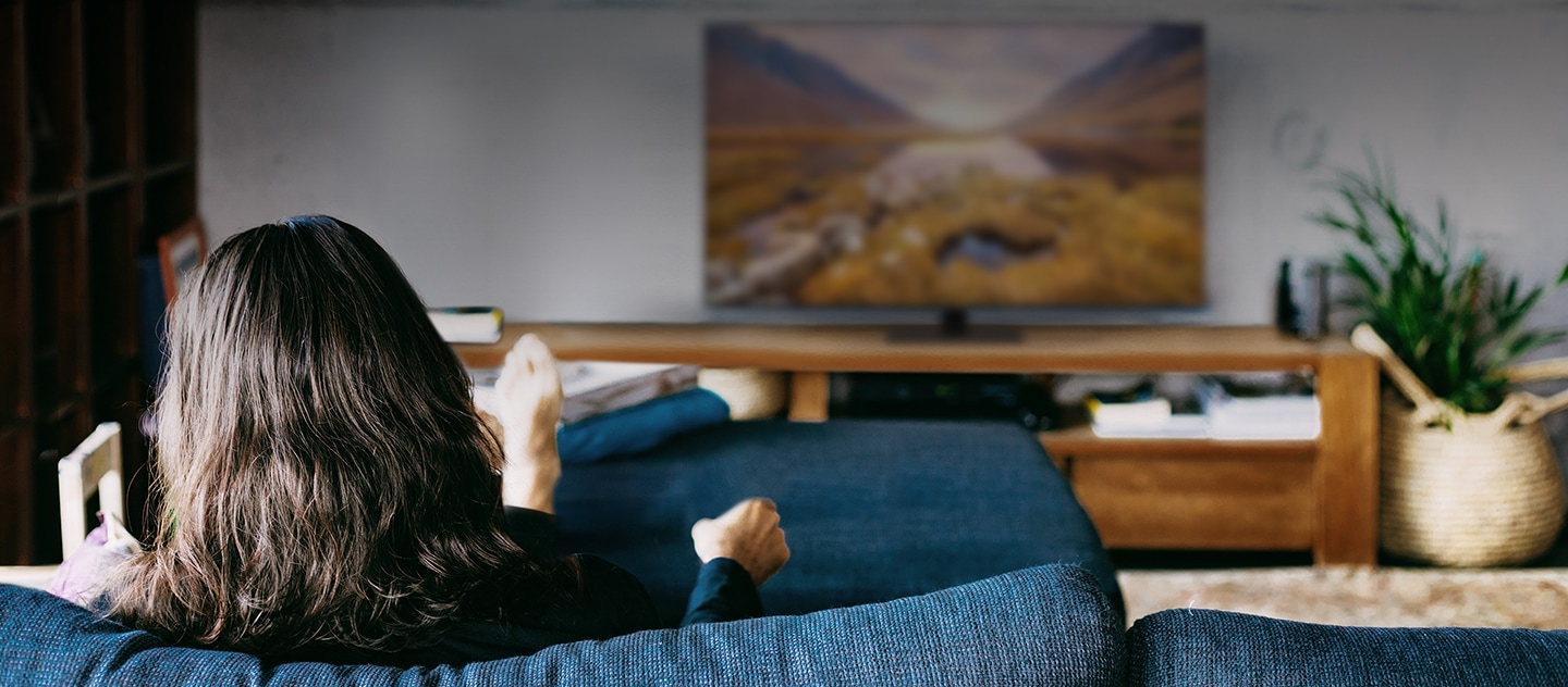 A woman, sitting on a sofa in her living room, is watching her living room TV. 