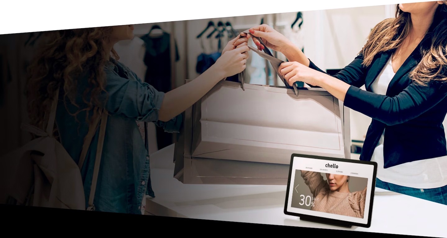 A customer at a clothing store is receiving her purchases from the store clerk. On the table between them is a Galaxy tablet device showing the store's online catalogue.