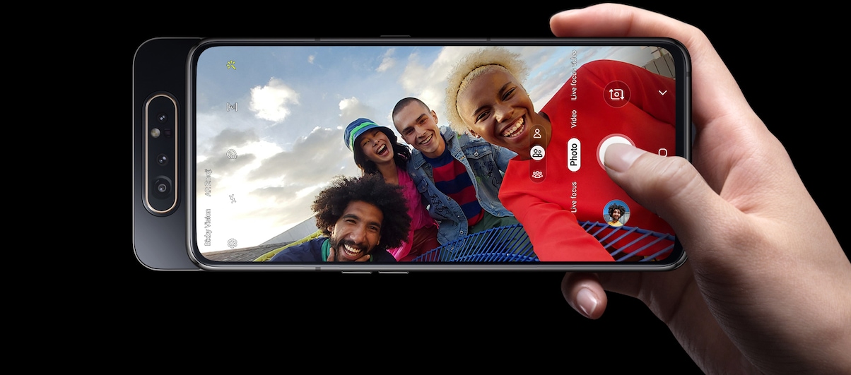 Celular Galaxy A80 sendo segurado na horizontal. Na tela é exibido o modo de fotografia. Na foto, um grupo de quatro amigos - duas mulheres e dois homens - sorri.