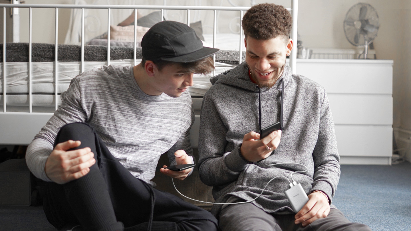 Foto de dois adolescentes sentados no chão de um quarto. Cada um está com um celular na mão e ambos os aparalhoes estão conectados ao carregador Samsung segurado por um deles.
