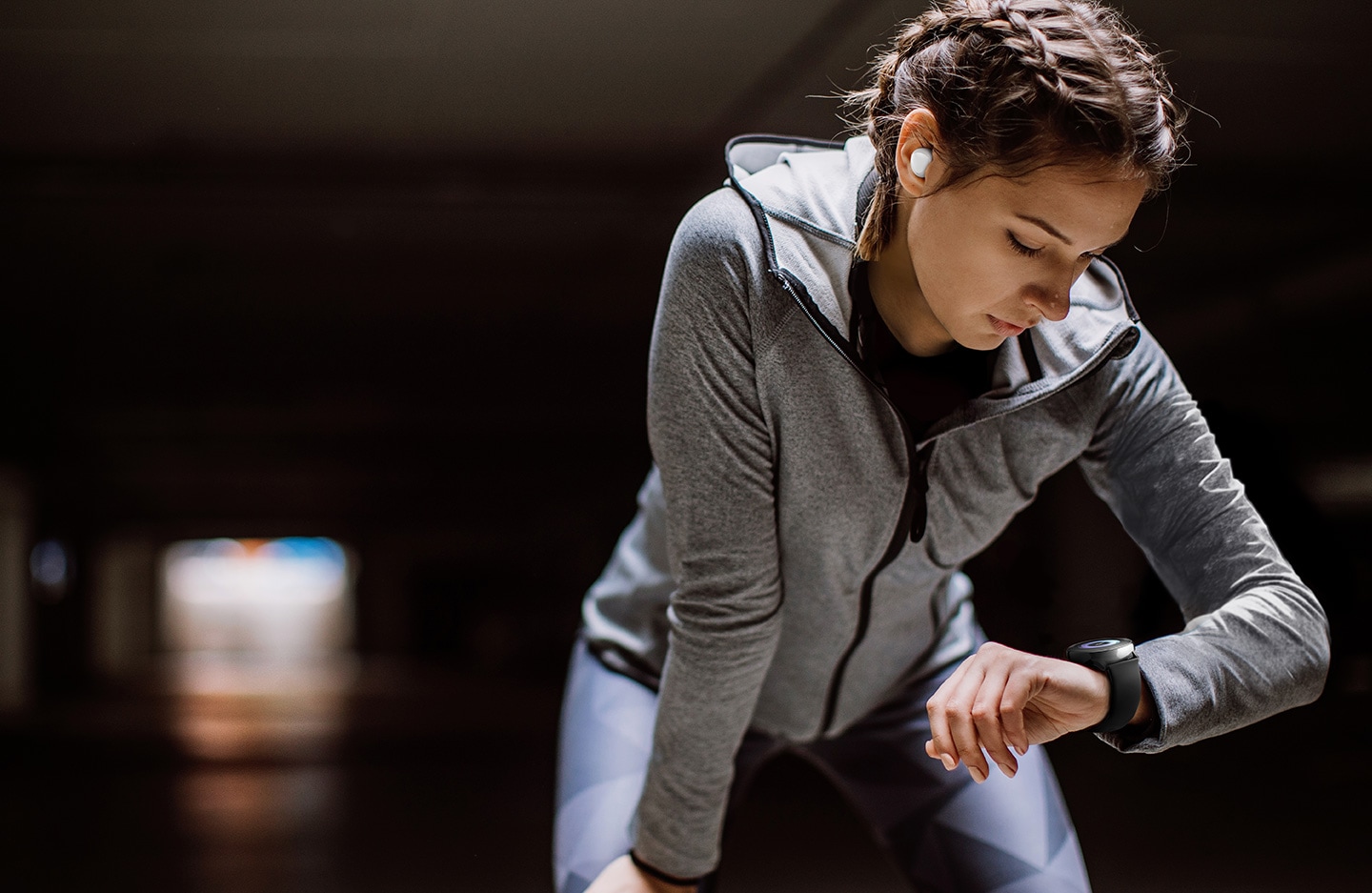 Uma mulher utilizando roupas esportivas flexiona levemente seus joelhos e olha para seu smartwatch Galaxy Watch Active no pulso esquerdo. Na orelha direita dela, é possível visualizar um Galaxy Bud branco.