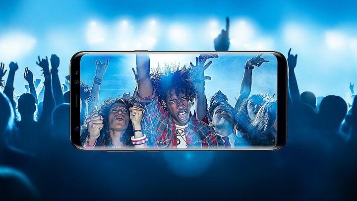 Ao fundo, ambiente com muitas pessoas de costas, pulando em uma festa. À frente, Galaxy S8+ deitado, com a tela na câmera, mostra as pessoas de frente, dançando na festa.