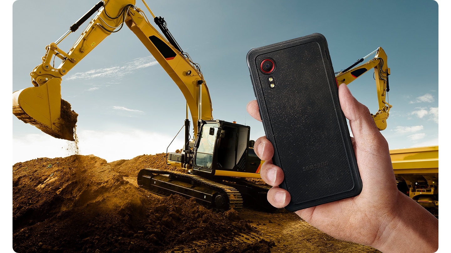 One hand holds the Galaxy XCover 5 in black and shows a dusty back.  In the background, yellow excavators are digging up earth on a construction site.