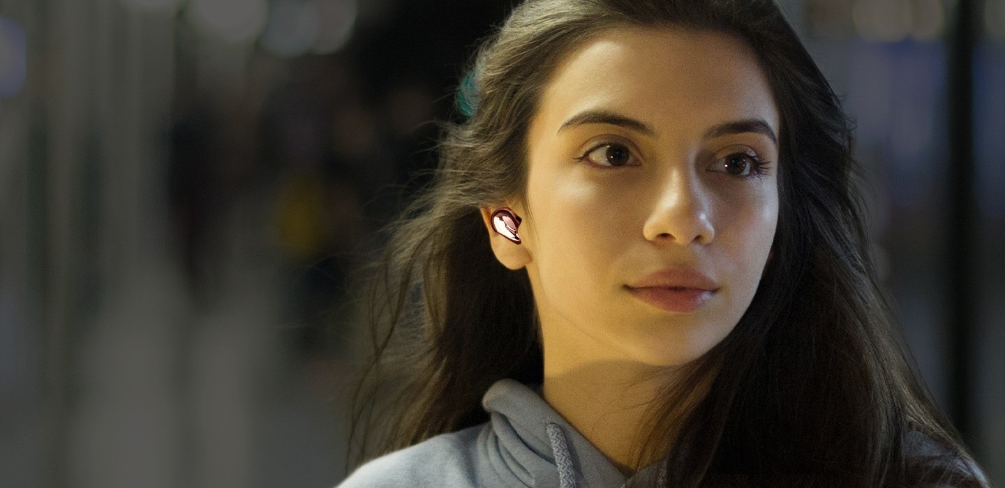 Woman standing on a busy subway platform wearing Galaxy Buds Live in Mystic Bronze with ANC off. A strong sound wave is shown underneath. Then as ANC is turned on, a close-up of the woman is seen with a dark and faded background, showing the effects of noise cancellation