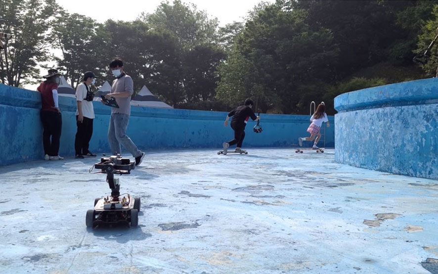 A mobile remote controlled unit carrying a Galaxy S20 Ultra as it follows an actor on a longboard through an empty pool. Three crew members stand nearby. 