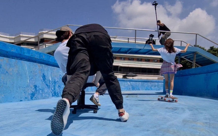 One crew member pushing a cameraman as he sits in a rolling office chair filming the actor with a Galaxy S20 Ultra as she longboards through an empty pool.