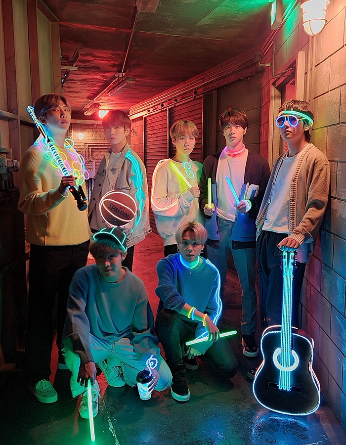 Photos taken with Night mode on Galaxy S20 plus. BTS stands in a low-lit hallway, holding objects lit up with glowsticks and neon lighting like a guitar, clarinet, basketball, sunglasses, and more. In the Night mode off photo, you can see outlines of the band members but mostly the glowsticks and neon objects. In the Night mode on photo, the faces and outfits of the band are much clearer and more colorful