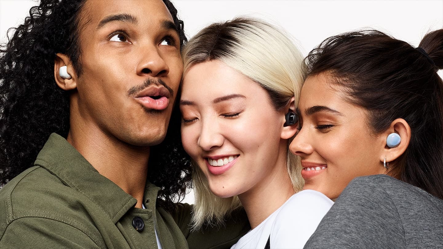 A close-up of three friends in a tight hug. A man on the left is wearing white earbuds and looks up while speaking. A woman in the middle wearing black earbuds closes her eyes and smiles. A woman on the right wearing blue earbuds rests her head on the middle woman's shoulder and smiles with her eyes closed. The Ambient sound volume GUI on the top right is turned on to the loudest setting.