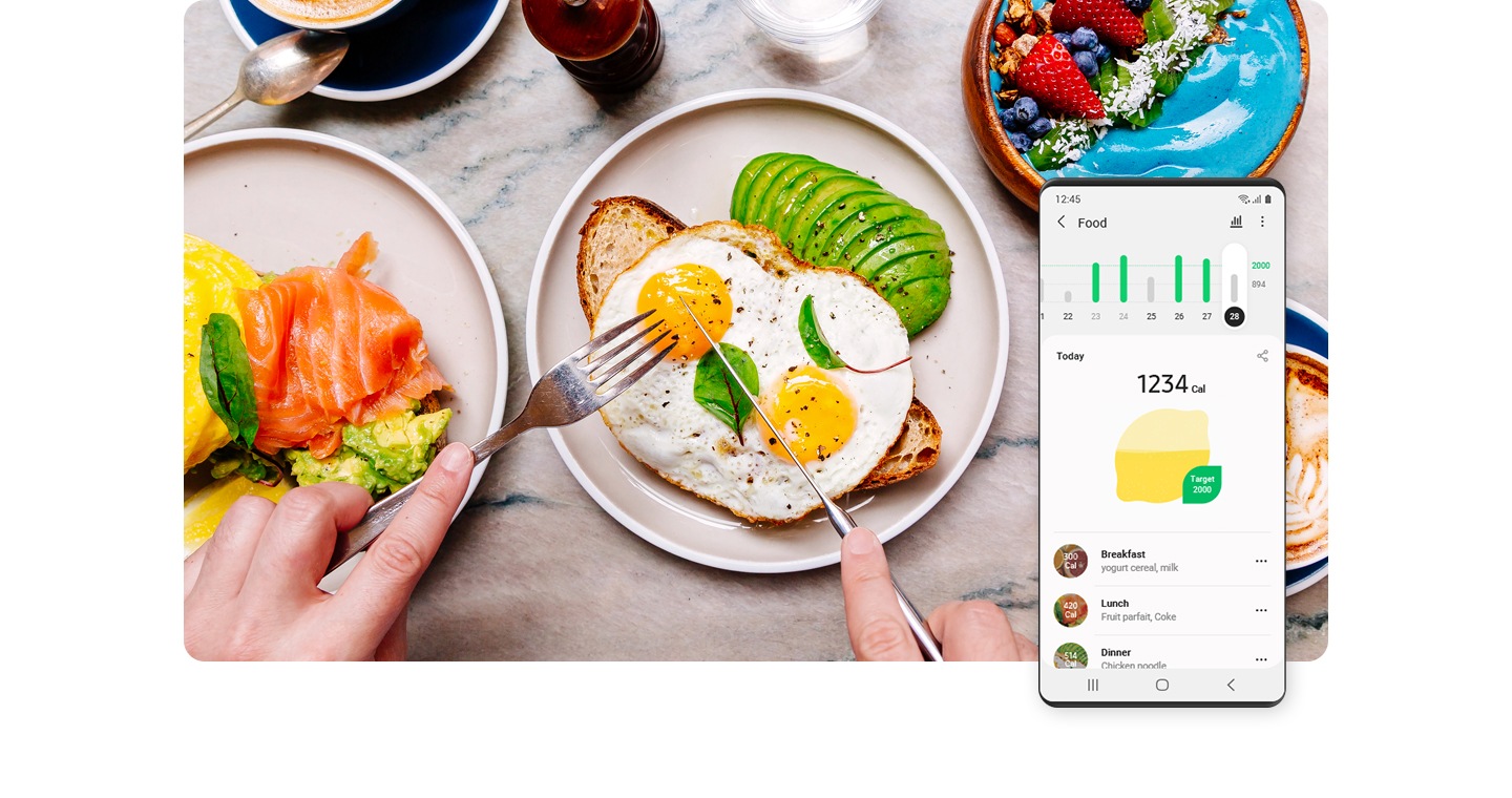 Galaxy smartphone is shown displaying the day's nutrition calorie count. Behind the smartphone, a breakfast containing eggs, toast and avocado is shown surrounded by a berry cake, coffee and a plate with salmon, eggs benedict and avocado toast.