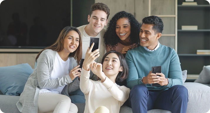 Een groepje vrienden zit op de bank terwijl de vrouw in het midden op de grond zit en haar smartphone omhoog houdt om een selfie van de groep te maken.