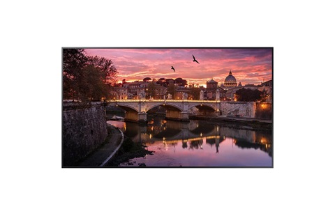 A Samsung monitor with a sunset, purple, pink, and yellow sky. There are birds flying in the sky. There is a skyline in the background, trees and a bridge with lights stretching from one end to the other end. Under the bridge is water that has a reflection of the bridge and sky.