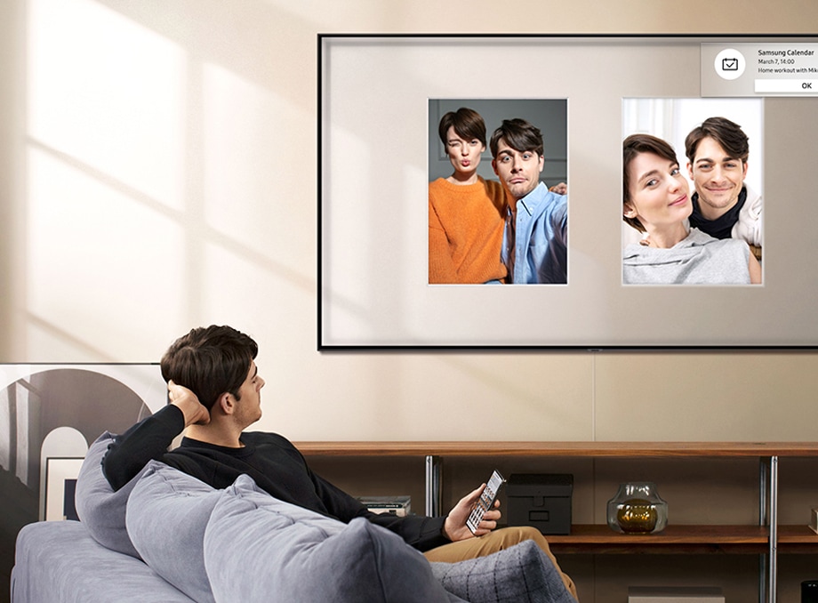 Un hombre está sentado en un sofá mirando un televisor con pantalla grande en casa y recibe notificaciones de la agenda directamente en la pantalla del televisor. 