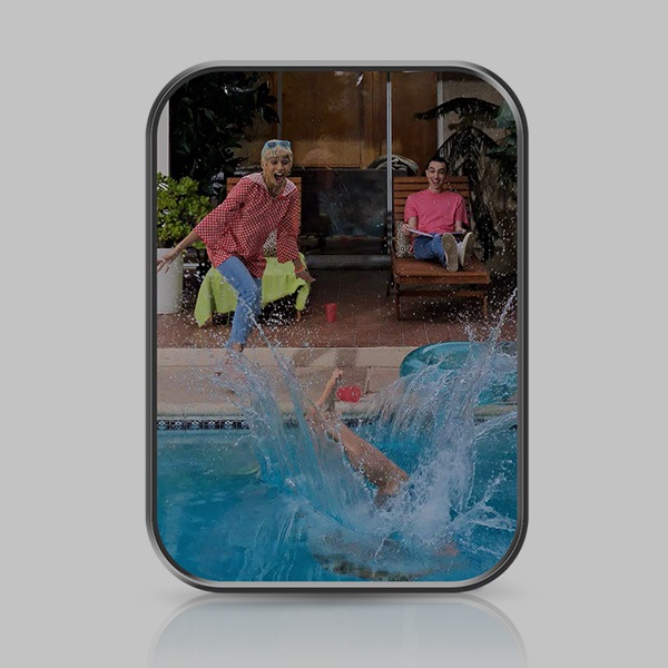 Una persona sumergida a medias y salpicando en una piscina al aire libre. Una mujer afuera de la piscina mira con la boca entreabierta. Un hombre también observa la escena mientras está sentado en una silla de la piscina junto a la mujer con un libro en su regazo.