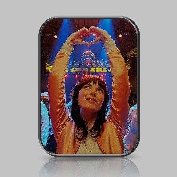 Una mujer realiza una forma de corazón con las manos en el aire. Dos reflectores se iluminan en el fondo. Se observa una bola de disco detrás de la mujer.