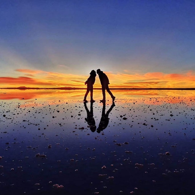 Una pareja parada sobre una superficie reflejada con un atardecer colorido en el fondo mientras posan enfrentados de forma divertida