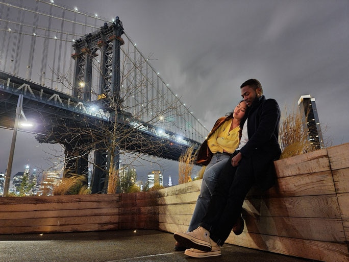 Una pareja sentada en una superficie de madera con el puente de Manhattan en el fondo; la escena se toma desde un ángulo bajo.