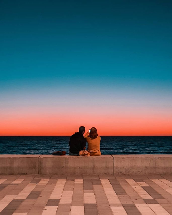 Una pareja sentada en una pared de concreto mirando hacia el océano. Se observa un atardecer impresionante que crea degradado colorido en el cielo.