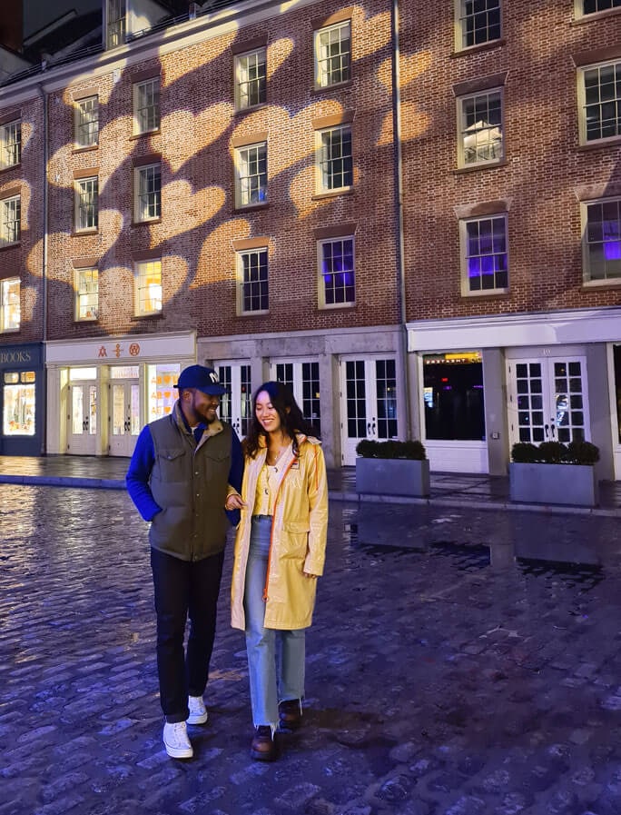 Una pareja que sonríe con los brazos enlazados y camina por una calle adoquinada. Hay luces con forma de corazón proyectadas en los edificios en el fondo.