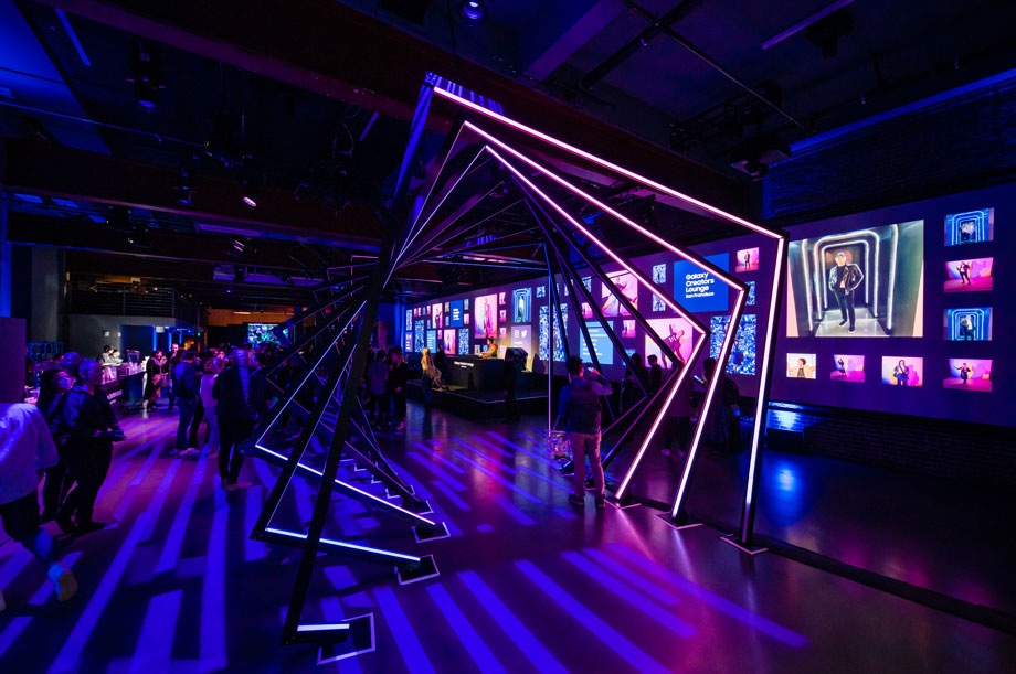 Influencers reunidos en el espacio de eventos del salón de los creadores Creators Lounge. Se puede ver una colorida instalación de luces en la parte delantera junto a un escenario para DJ y un bar en la parte trasera.
