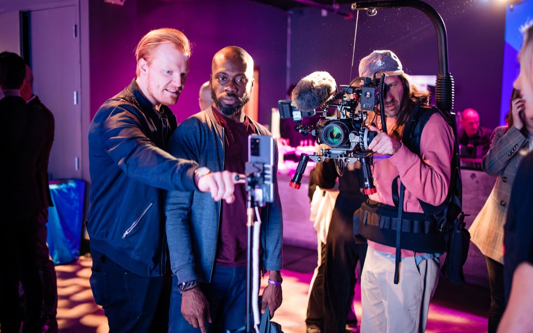 Dos hombres están de pie junto a un Galaxy S20 Ultra 5G colocado en un soporte. Uno de los hombres está tocando la pantalla mientras un camarógrafo filma la escena a su lado.