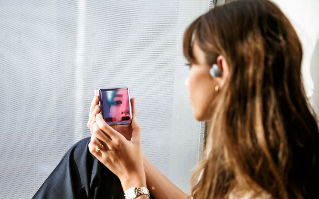 Una vista de perfil lateral de una mujer sentada. Tiene un auricular Galaxy Buds plus en el oído y con las manos sostiene el teléfono Galaxy Z Flip frente a ella.