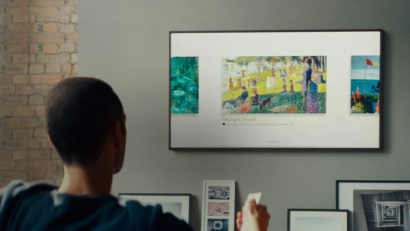Muž používa diaľkové ovládanie na prehliadanie niektorých obrazov, ktoré sú zobrazené na The Frame TV, ktorá je pripevnená na stene v obývačke.