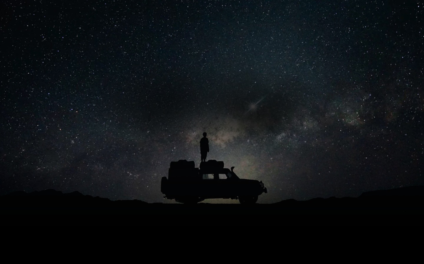 En person, der står oven på sin bil om natten, ses mod stjernehimlen