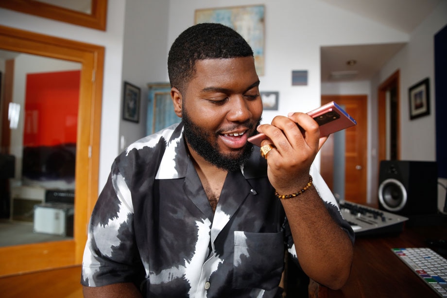 خالد يحمل الهاتف Galaxy Note20 باللون البرونزي ميستيك ويضعه أمام فمه ويستخدم ميزة تحويل الكلام إلى نص في استوديو التسجيل. إنه يرتدي قميصًا بأزرار مصبوغًا باللونين الأبيض والأسود.