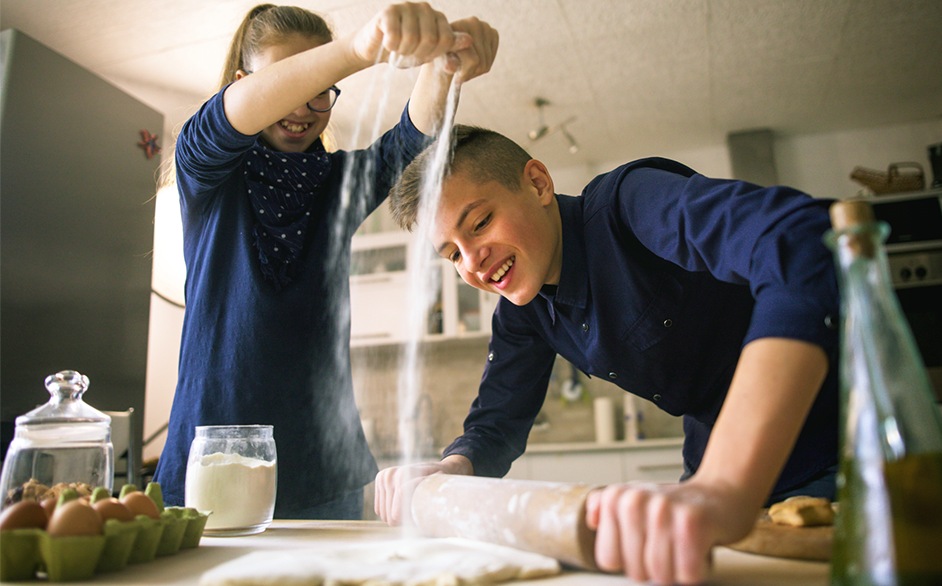 The brother and sister smile as they roll out their bread dough on the kitchen counter. He’s  using a wooden roller as she gradually adds more flour. 