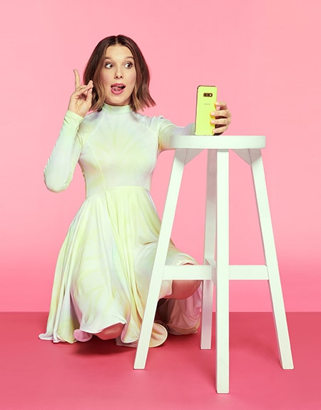 Millie Bobby Brown kneels while holding up a peace sign with her fingers as she takes a selfie with a neon yellow Samsung Galaxy A80 that's propped on a white stool