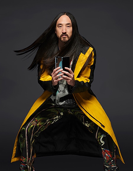 Steve Aoki standing with his knees slightly bent outward while sporting a yellow long coat as he holds up an unfolded Samsung Galaxy Fold smartphone.
