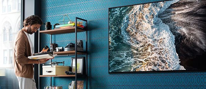 A man stands in front of a bookshelf in a living room, as the QLED 8K hangs on the wall, featuring an image of ocean waves. 