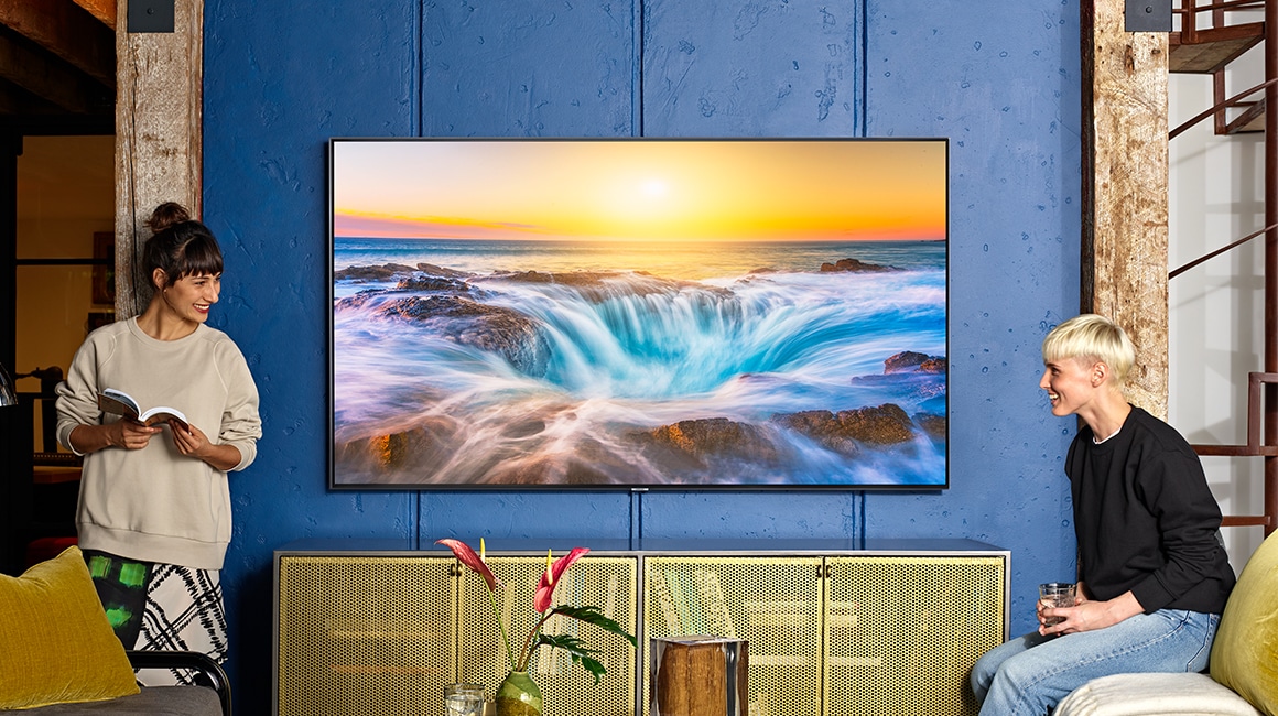 Una imagen lifestyle del nuevo Samsung QLED Q85R 2019. La imagen muestra el modelo Q85R montado en una pared azul, mostrando un aspecto visual muy limpio. Dos jóvenes hablan entre ellos frente al televisor.