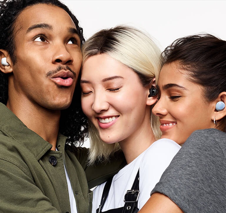 A close-up of three friends in a tight hug. A man on the left is wearing white earbuds and looks up while speaking. A woman in the middle wearing black earbuds closes her eyes and smiles. A woman on the right wearing blue earbuds rests her head on the middle woman's shoulder and smiles with her eyes closed. The Ambient sound volume GUI is turned on to the loudest setting.