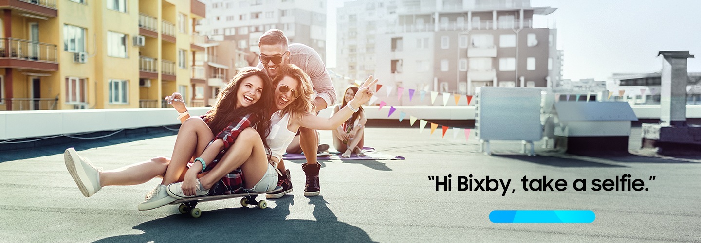 2 women sit on a skateboard and a man crouches behind them. A progress bar icon and text "Hi Bixby, take a selfie" are shown. 