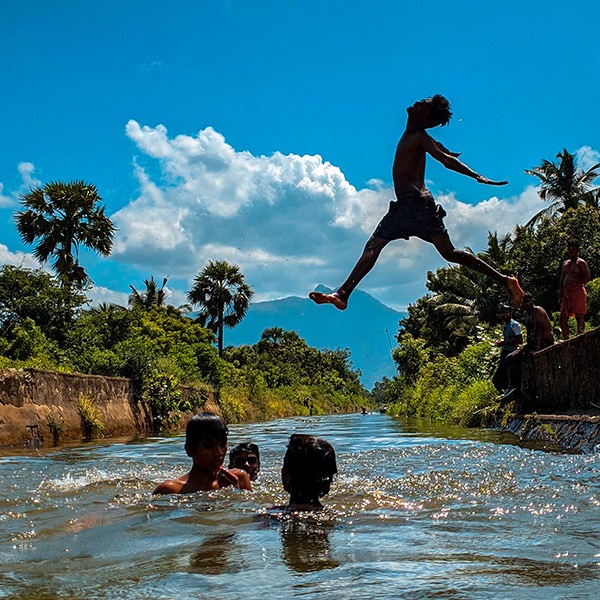 گروهی از مردان جوان در دریاچه شنا می‌کنند و یکی از آنها در حالی که درون آب می‌پرد، دستها و پاهایش را در هوا باز می‌کند